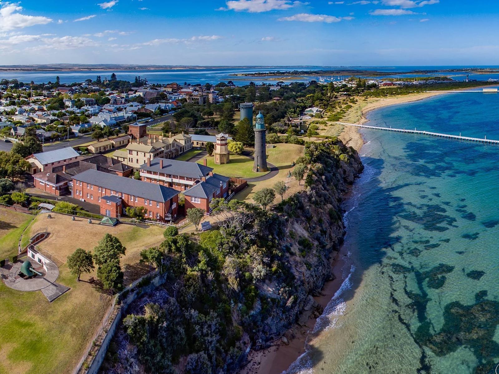 Aerial view of a coastal town featuring historic buildings, a lighthouse, and a sandy beach with clear blue waters.