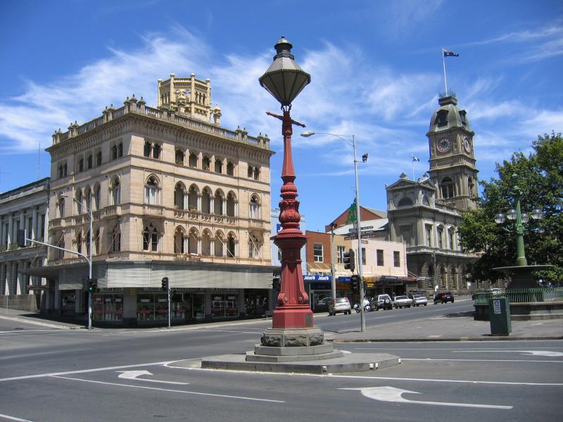A historic street view featuring ornate architecture, including a large building with a decorative facade and a clock tower, alongside a red street lamp and shops, set against a clear blue sky.