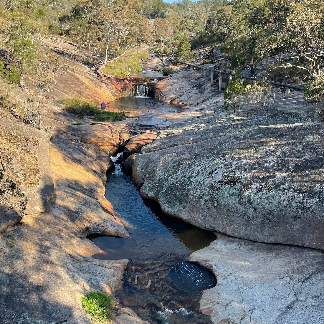 A serene landscape featuring a rocky stream with small waterfalls, surrounded by trees and greenery.