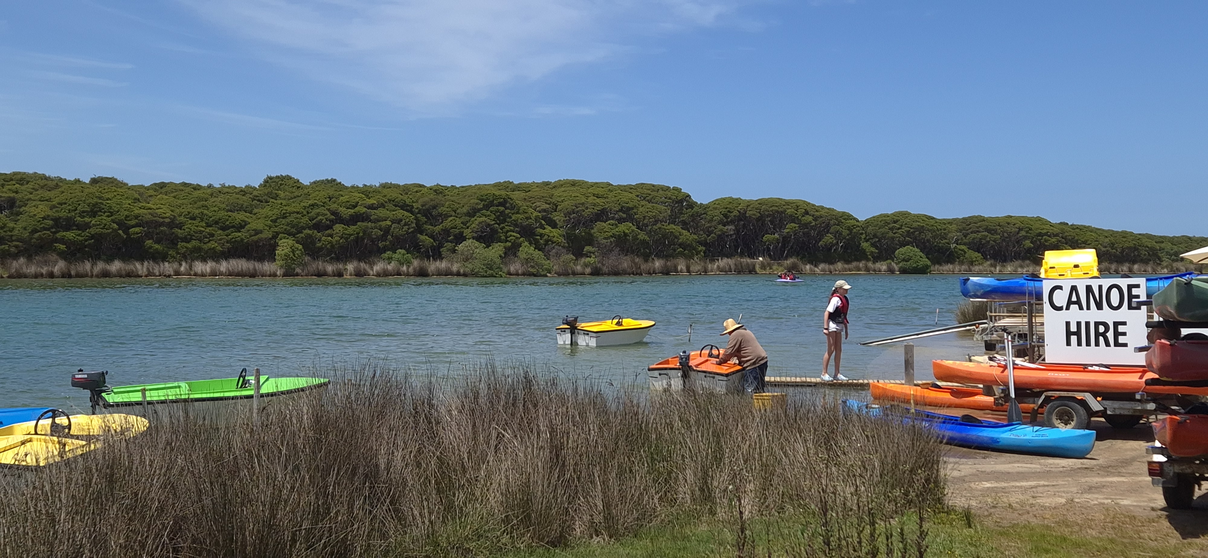A scenic view of a waterway with several colorful boats, including canoes and motorboats, docked near the shore. Two people are interacting with the boats, while a large sign reads 'CANOE HIRE'. Lush green trees line the background under a clear blue sky.