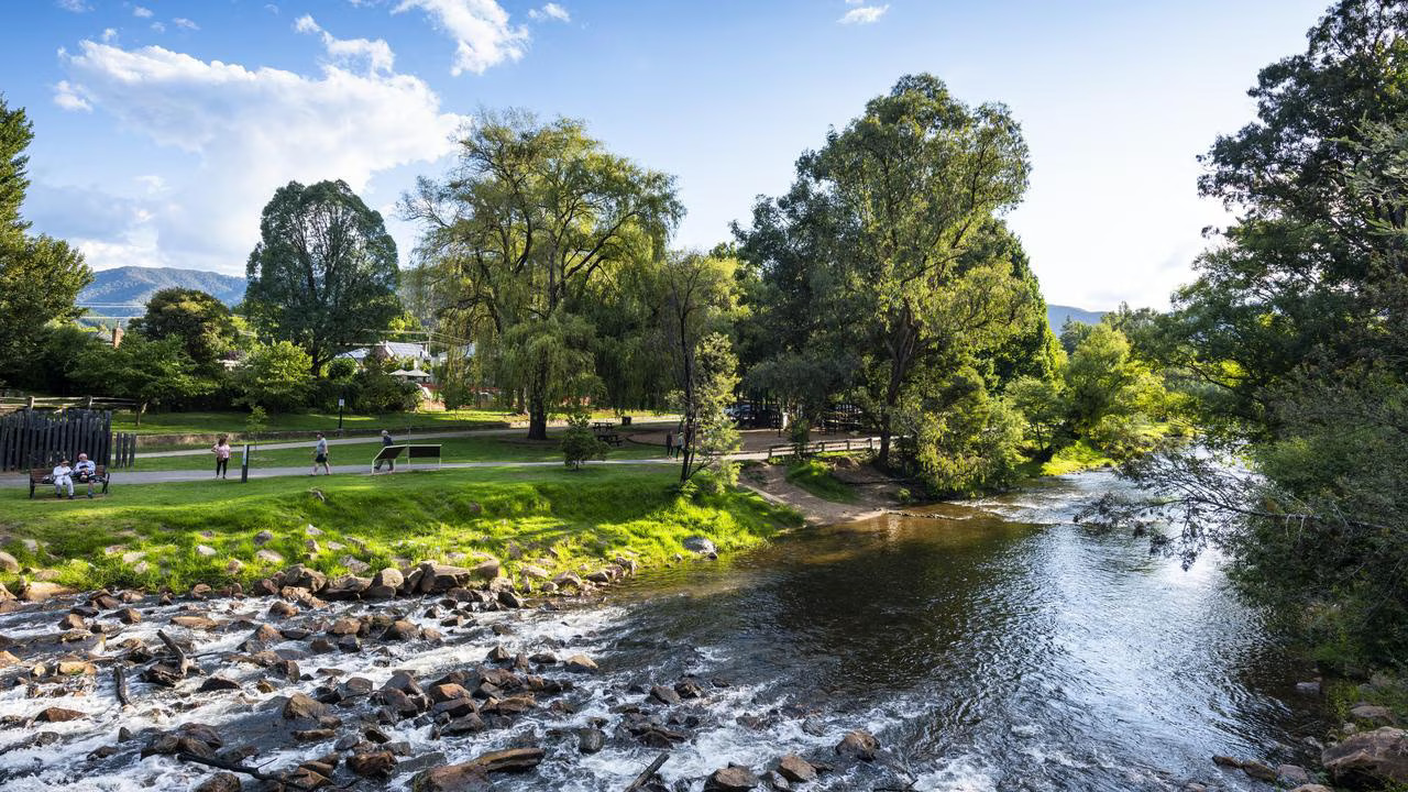 A scenic view of a river flowing through a green landscape with trees and pathways, where people are walking and enjoying the outdoors.