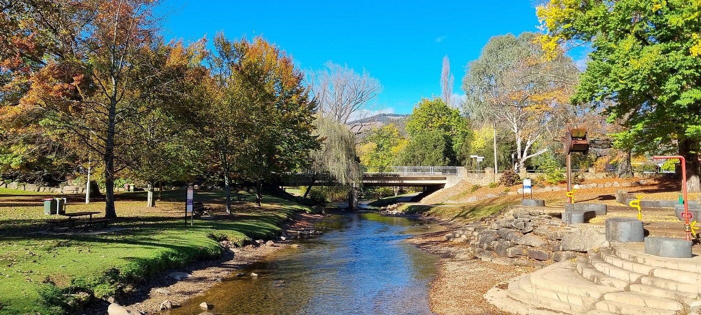 A serene park scene featuring a stream running through it, surrounded by trees in autumn colors. A bridge is visible in the background, and there are benches and play equipment near the water's edge.