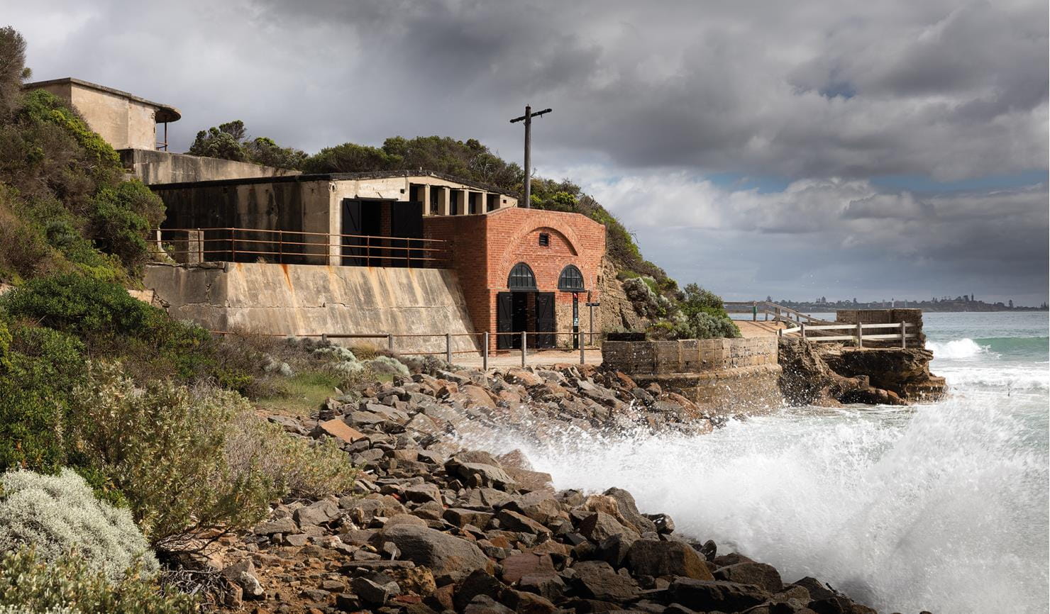 An old brick building situated on rocky shore with crashing waves and lush greenery surrounding it under a cloudy sky.