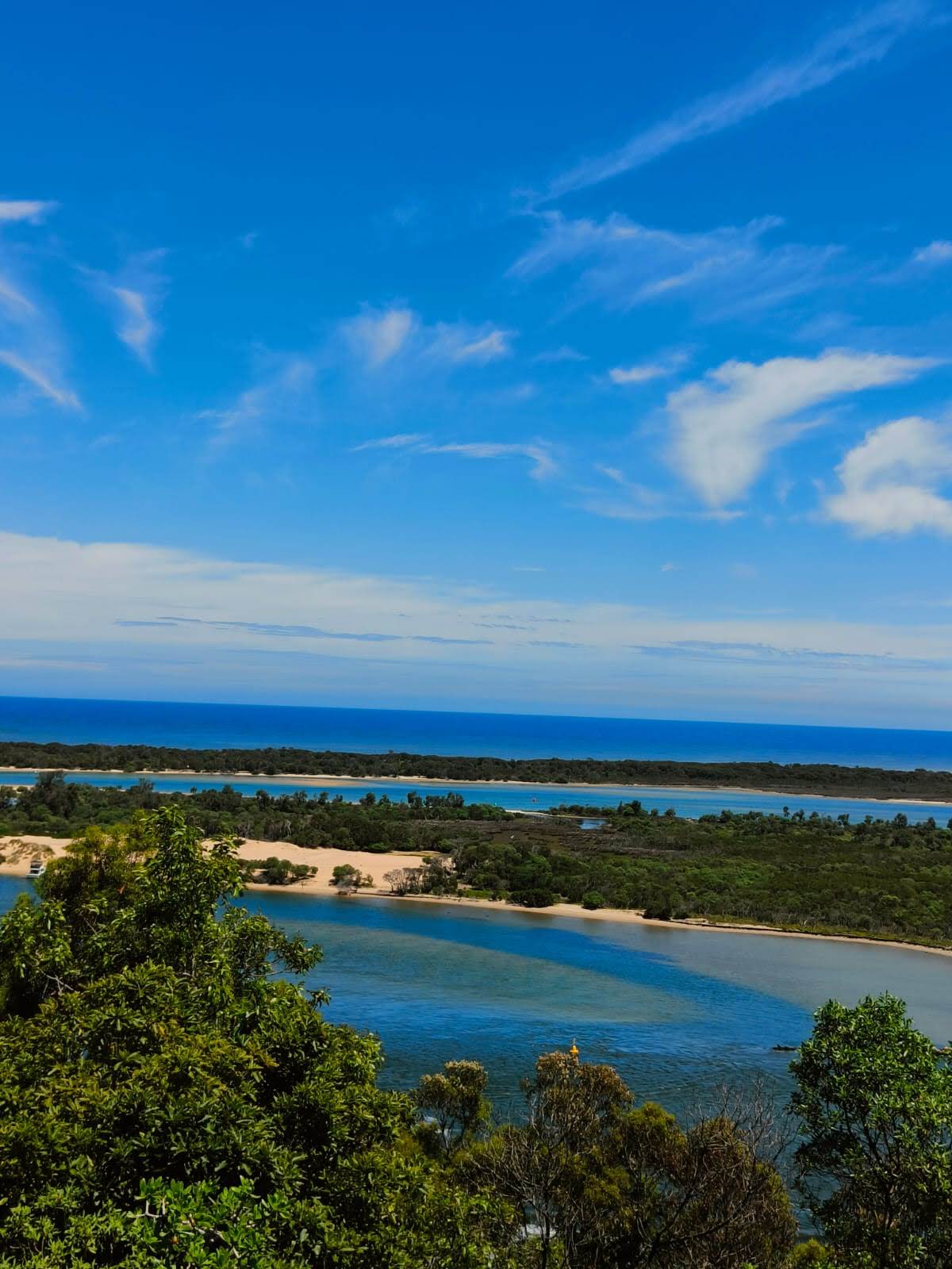 A scenic view of a coastline featuring blue waters and sandy beaches, framed by lush greenery under a clear blue sky with scattered clouds.