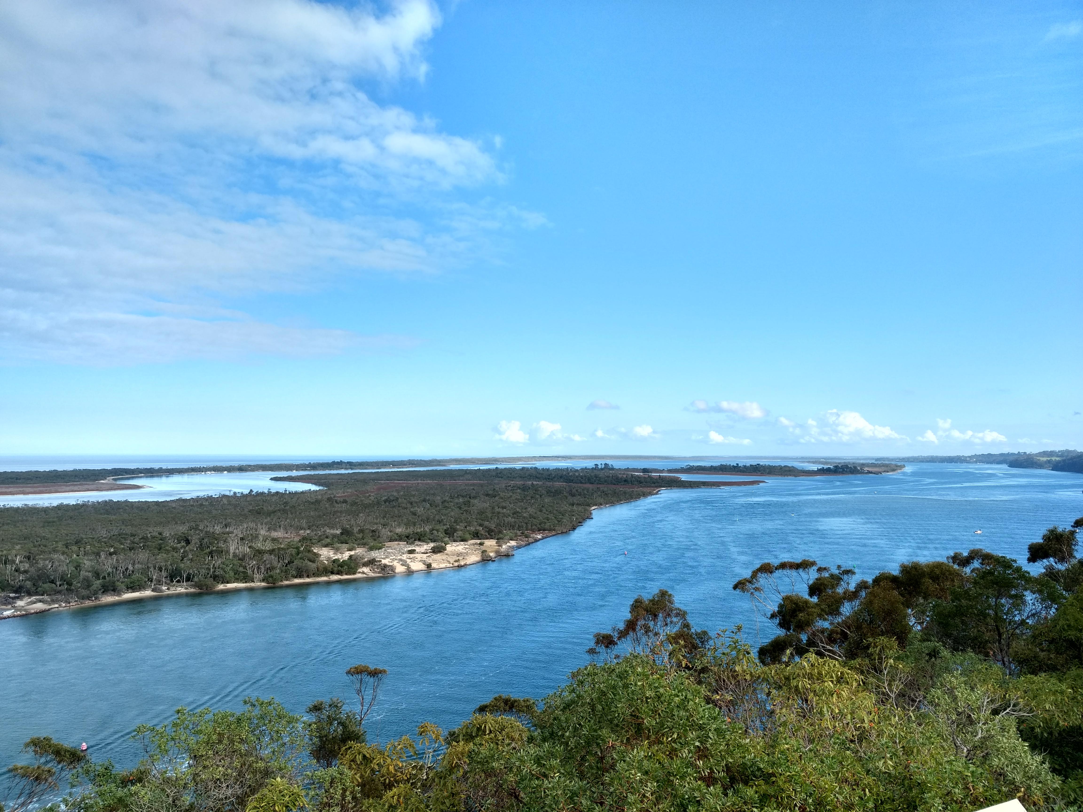 A panoramic view of a river meandering through lush greenery under a clear blue sky with scattered clouds.
