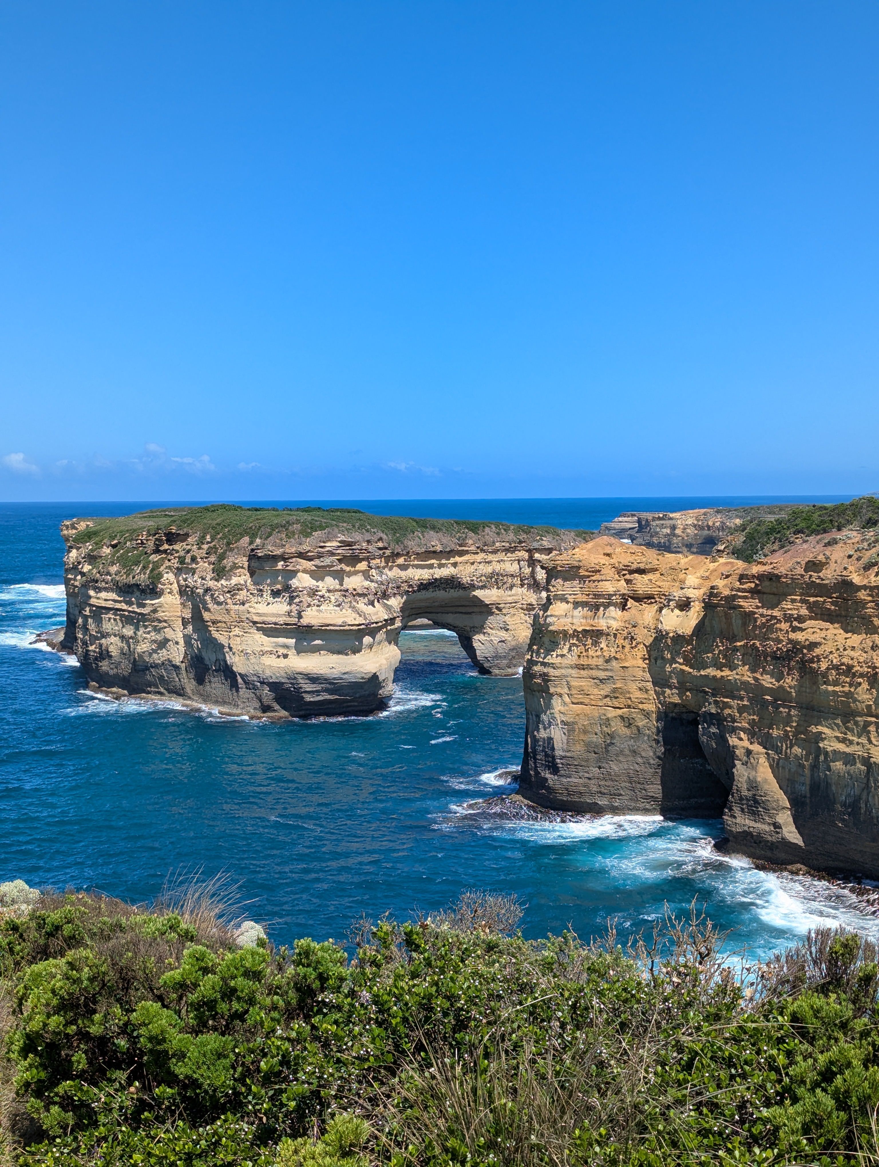 Scenic view of a natural rock arch over ocean water, surrounded by cliffs and greenery under a clear blue sky.