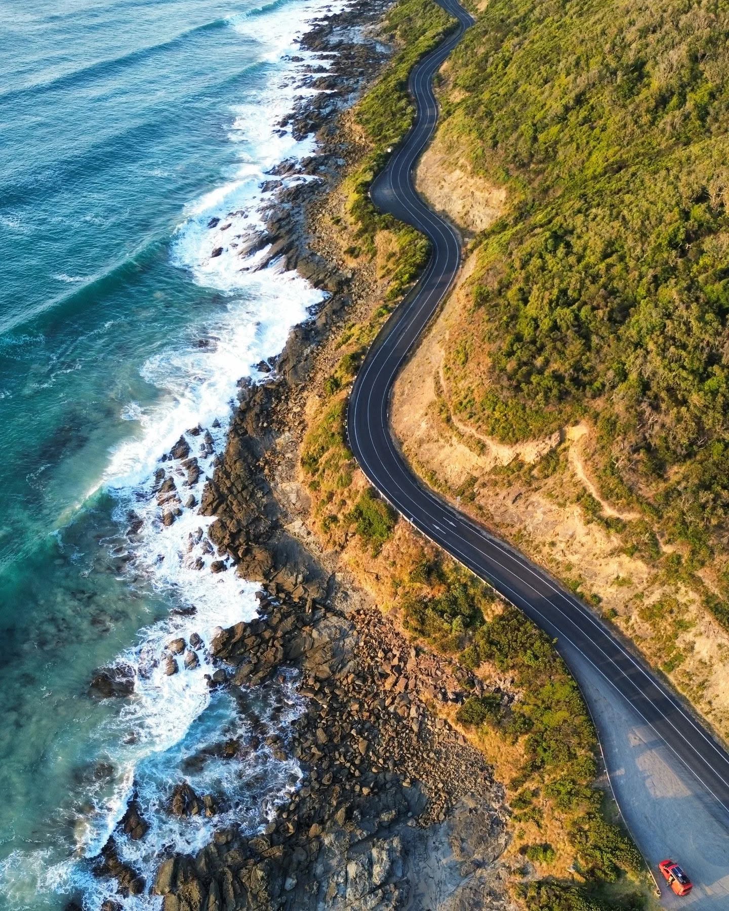 Aerial view of a winding coastal road alongside rocky cliffs and waves crashing against the shore.