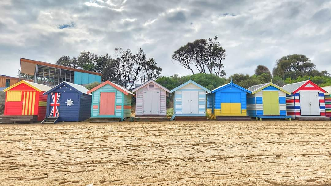 A row of colorful beach huts along a sandy shore, with a cloudy sky in the background.