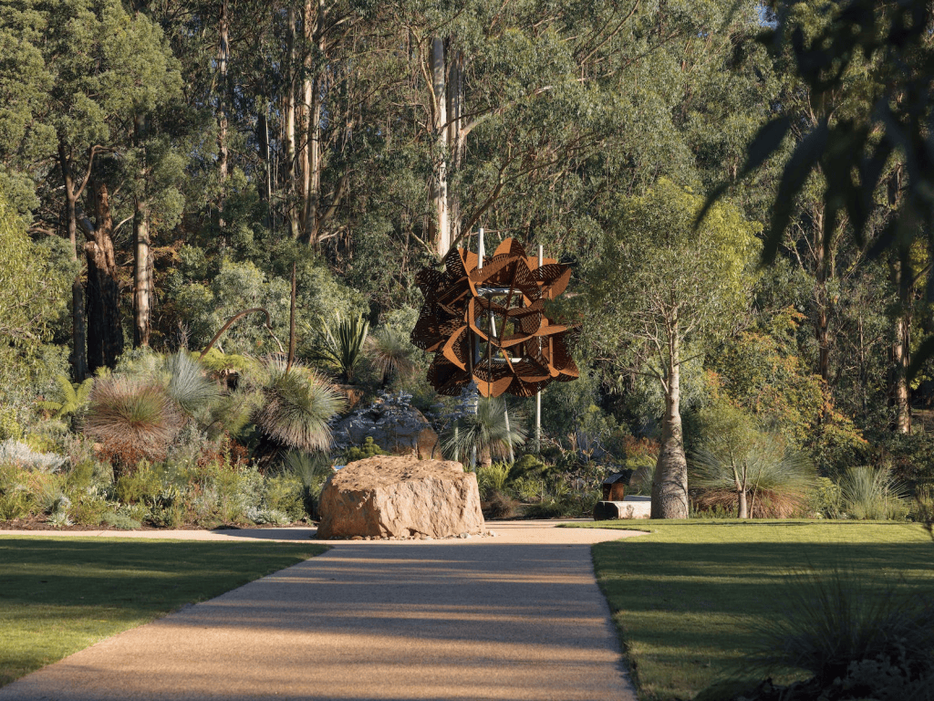 A landscaped garden featuring a winding path, surrounded by various plants and trees, with a large, abstract sculpture in the center.