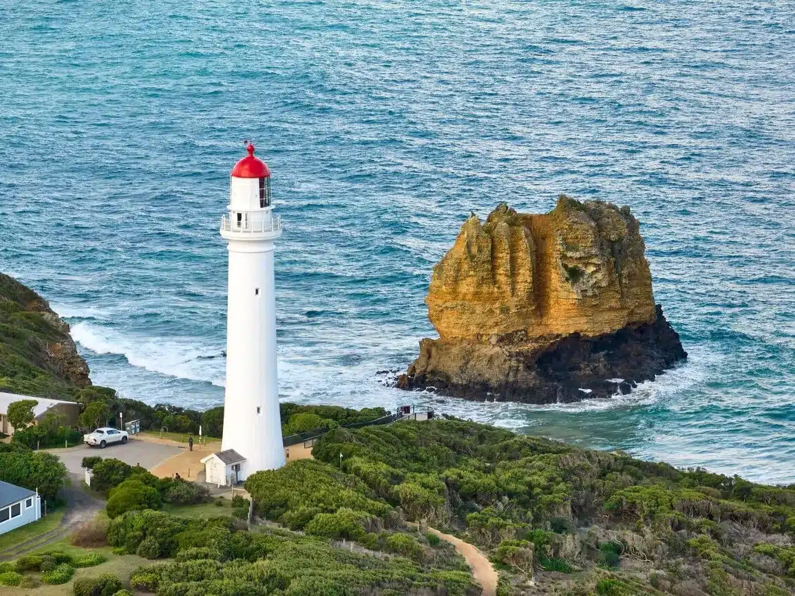 A tall white lighthouse with a red top stands near a rocky cliff, overlooking the ocean with gentle waves, surrounded by green vegetation.