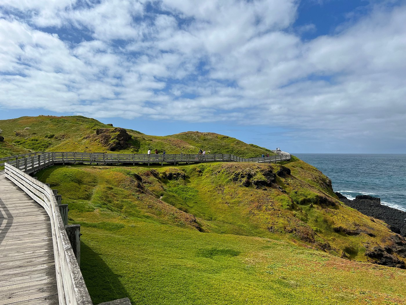 A scenic coastal view featuring a wooden boardwalk winding through lush green hills, with people walking along the path and the ocean visible to the right under a partly cloudy sky.