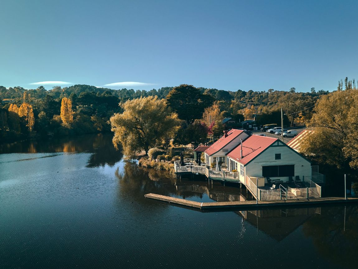 A serene lakeside view featuring a cozy white building with a red roof, surrounded by vibrant autumn foliage and reflecting on the calm water.