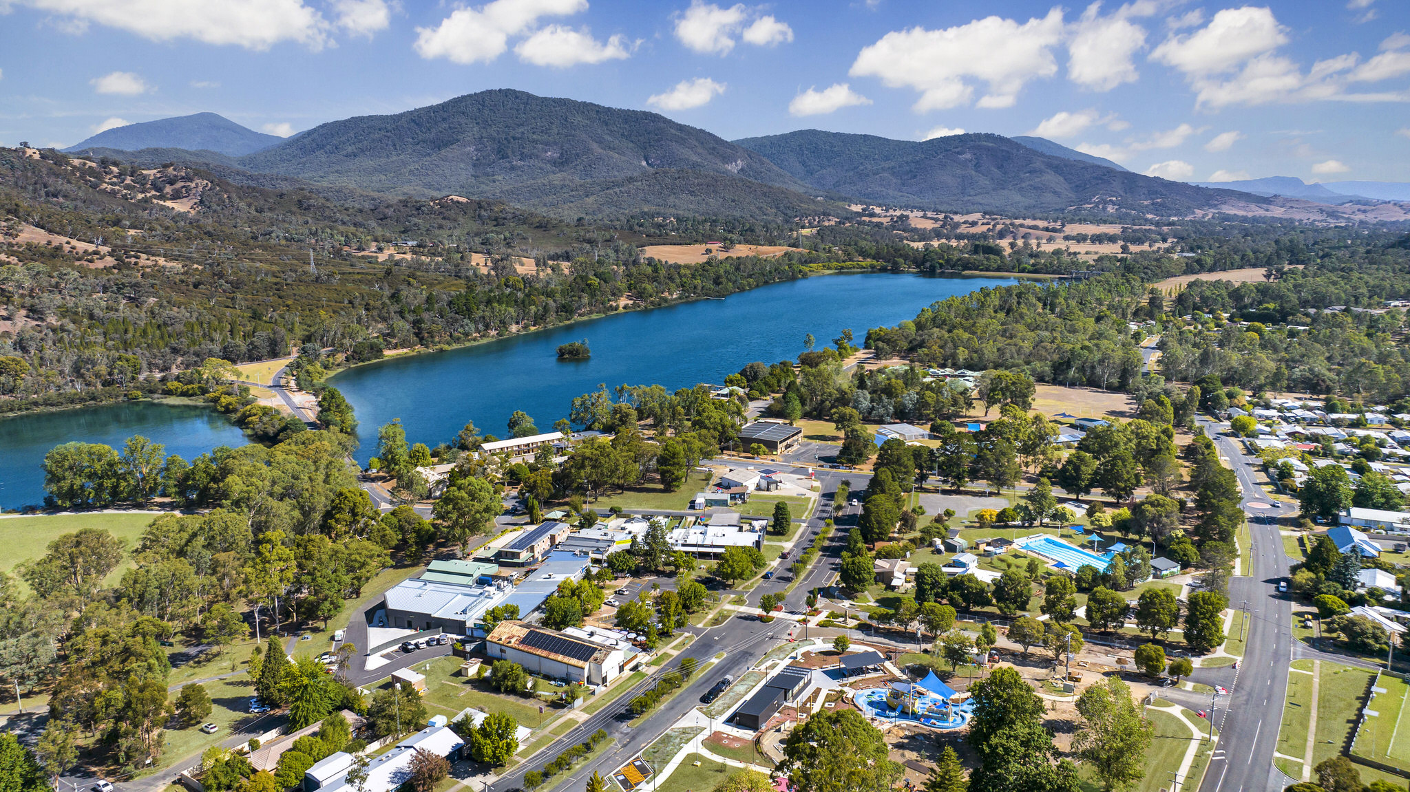 Aerial view of a river surrounded by lush greenery and hills, with a residential area and recreational facilities visible along the riverbank.