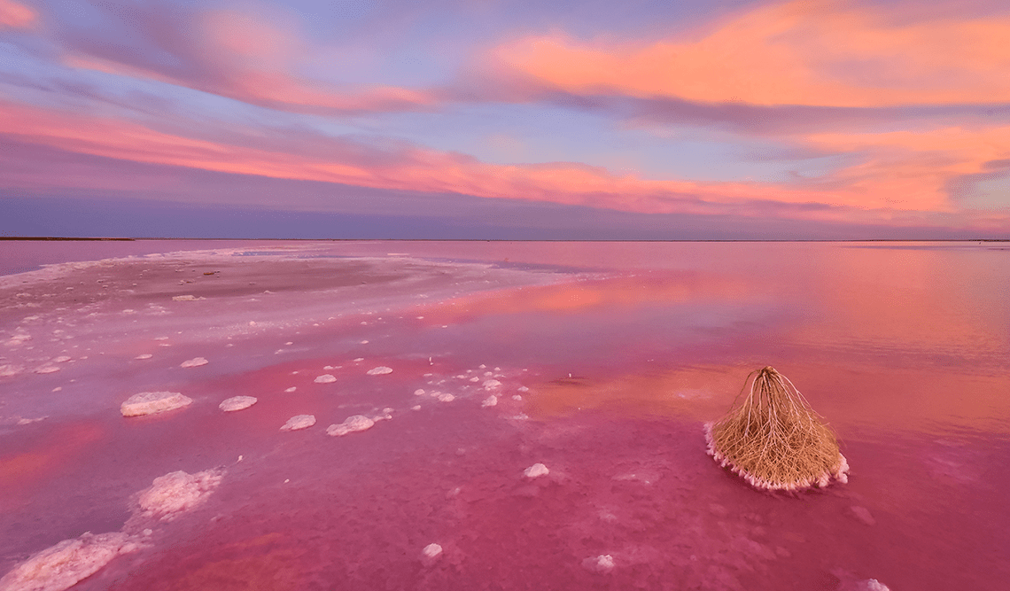 A scenic view of a pink salt lake during sunset, with colorful clouds reflecting in the water and a salt formation in the foreground.