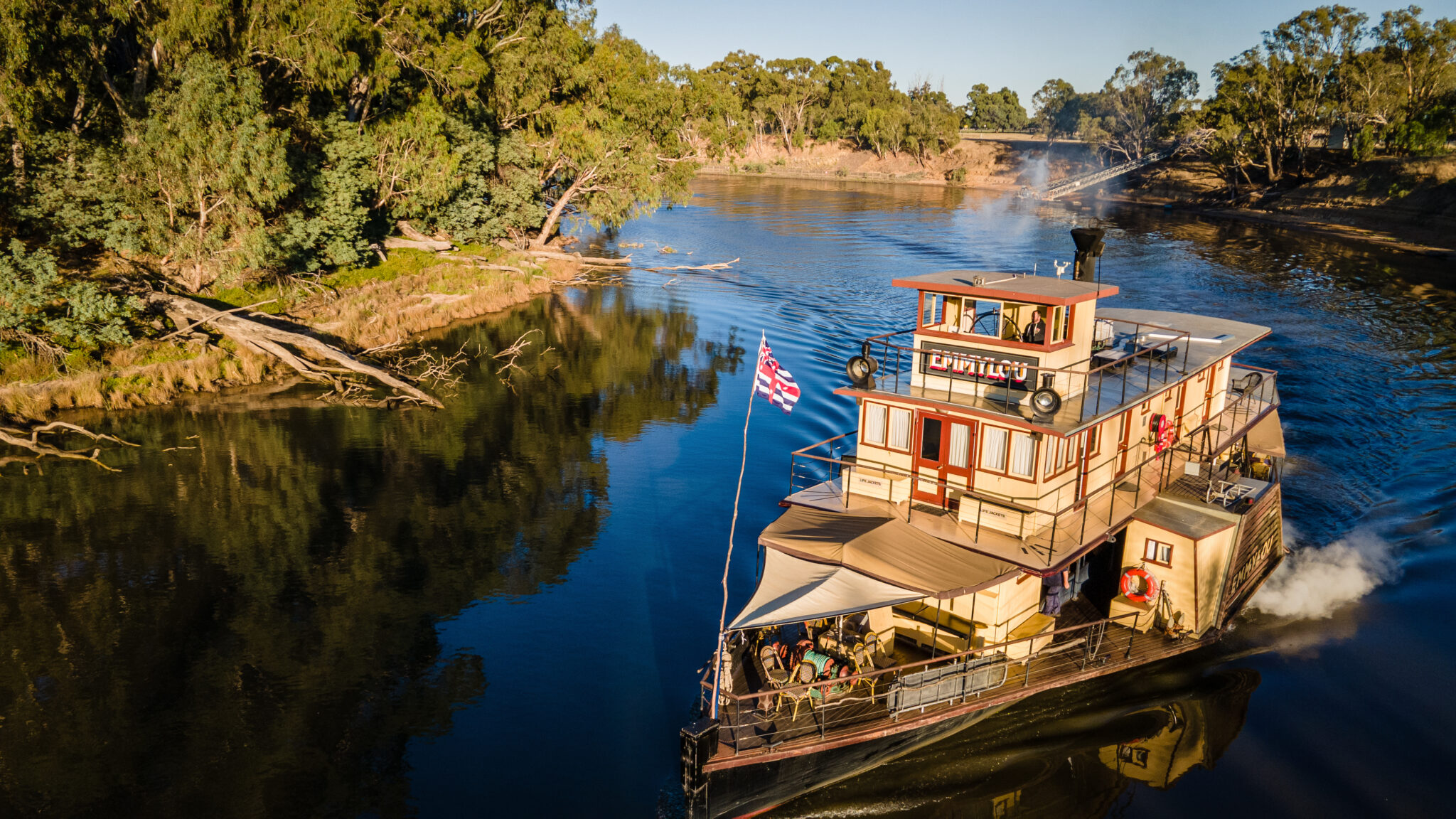 Aerial view of a vintage paddle steamer cruising on a river, surrounded by lush greenery and calm waters.