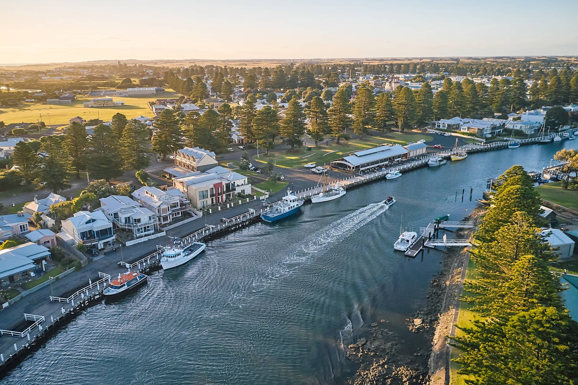 Aerial view of a tranquil river lined with houses and trees, featuring several boats navigating through the water.