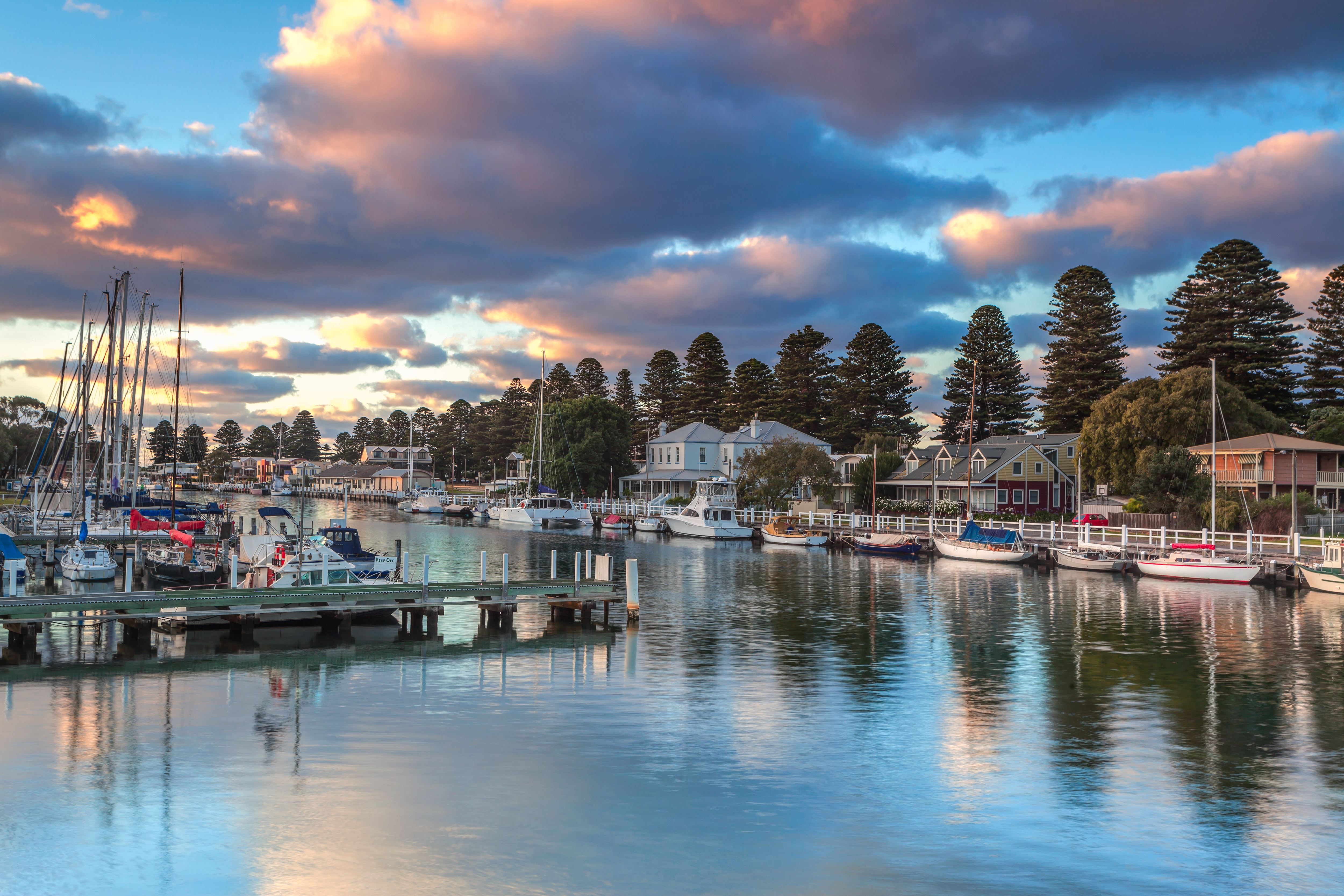 A serene waterfront view at sunset, featuring a marina filled with various boats and yachts, surrounded by lush trees and charming buildings reflecting in the calm water.