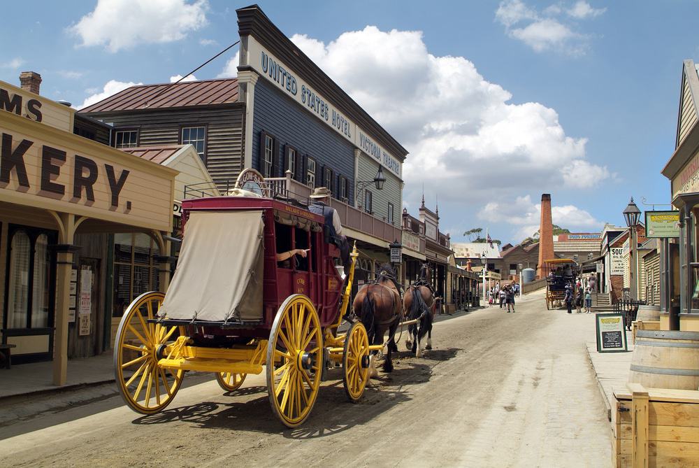 A vintage horse-drawn carriage on a dusty street in a historic town, surrounded by old-style buildings, shops, and clear blue skies.