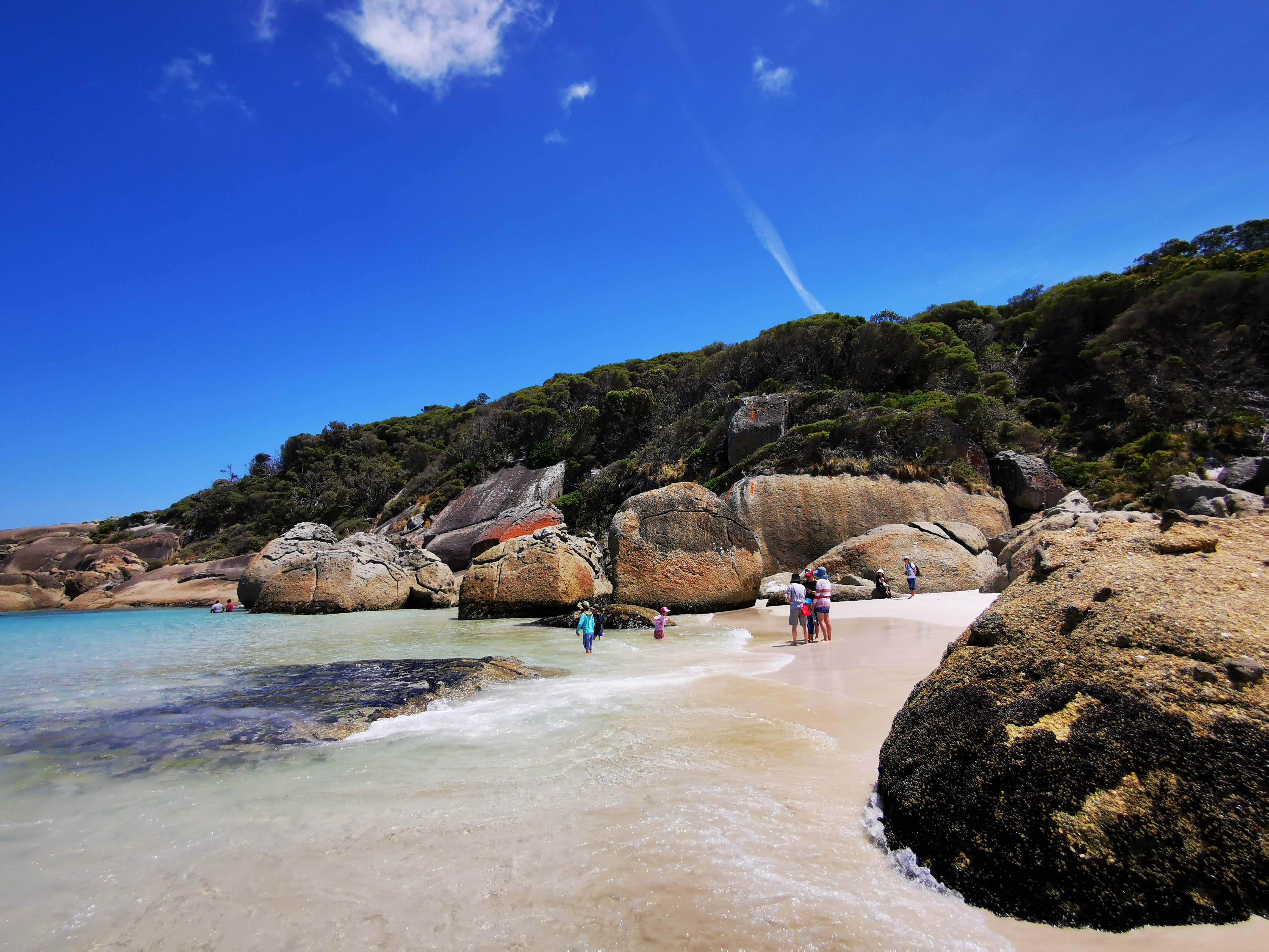 A scenic beach with large rocks and people exploring in shallow water under a clear blue sky.