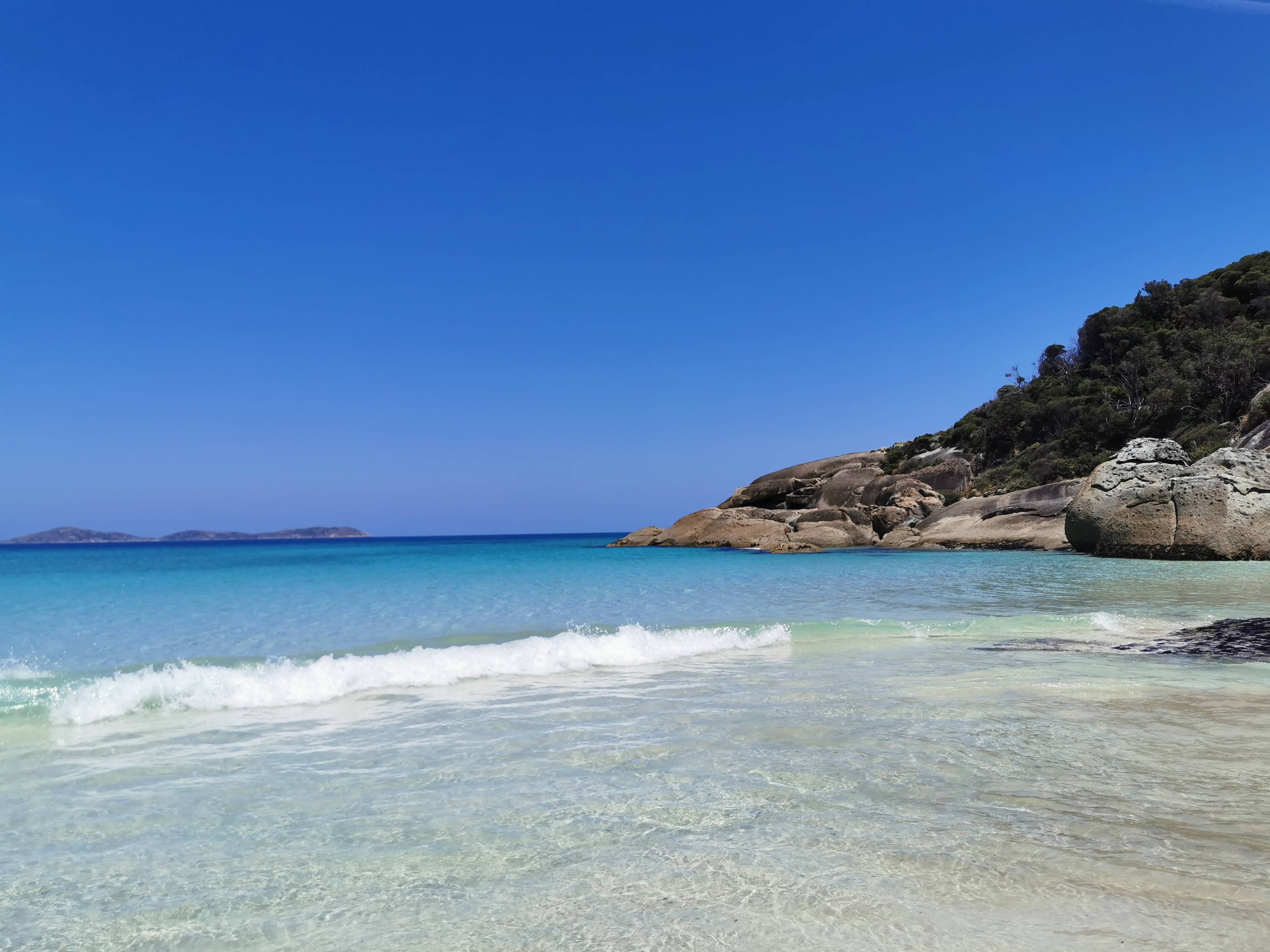 Calm beach scene with clear turquoise water, gentle waves, and rocky coastline under a blue sky.
